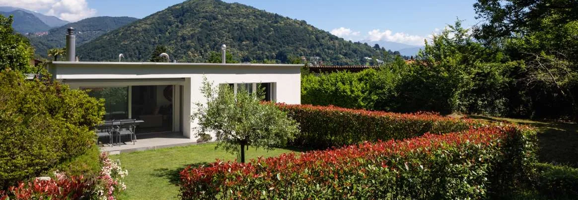 Modernes Haus mit Garten und Hecken vor einer Bergkulisse.